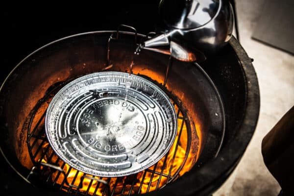 Víz öntése Big Green Egg alumínium csepegtető tálcába grillben, parázs fölött, ízes gőzös sütés előkészítése során

