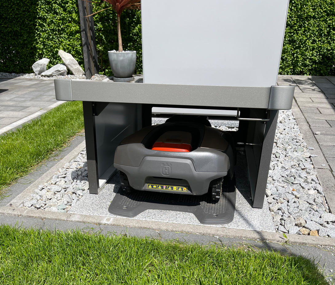 Biohort Charly robotfűnyíró garázs használat közben, benne egy Husqvarna robotfűnyíró töltőállomáson, modern térkövezett és kavicsos környezetben.