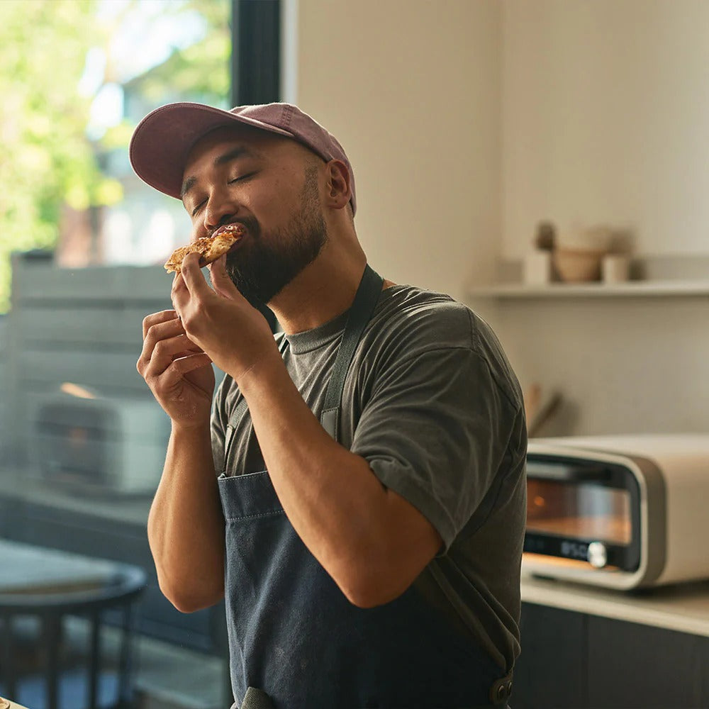 Felhasználó pizzaszeletet kóstol Ooni Volt 2 elektromos pizzasütő mellett, beltéri környezetben.