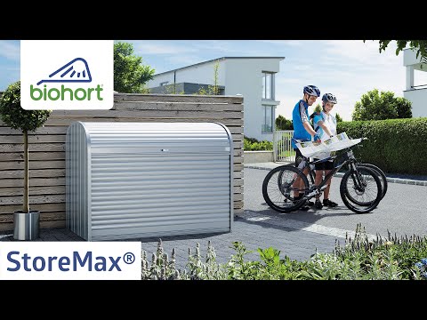 Biohort StoreMax tároló láda ezüstmetál színben, modern ház előtt elhelyezve, ideális kerékpáros kiegészítők és kerti eszközök kültéri tárolására.